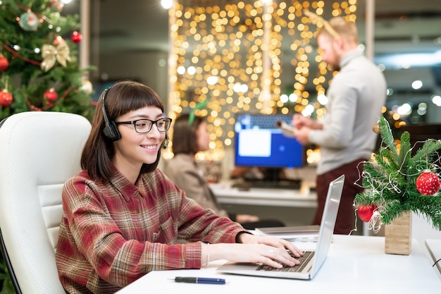 Pretty young brunette operator with headset typing in front of laptop while organizing work in office on xmas day