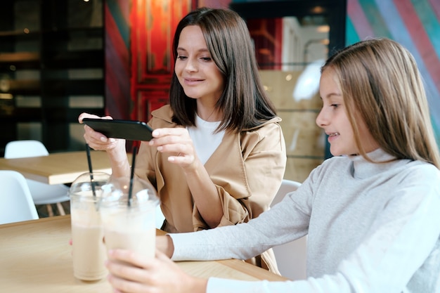 スマートフォンで娘と一緒に時間を過ごしながらカフェで2つのミルクカクテルの写真を撮るとかなり若いブルネットの女性