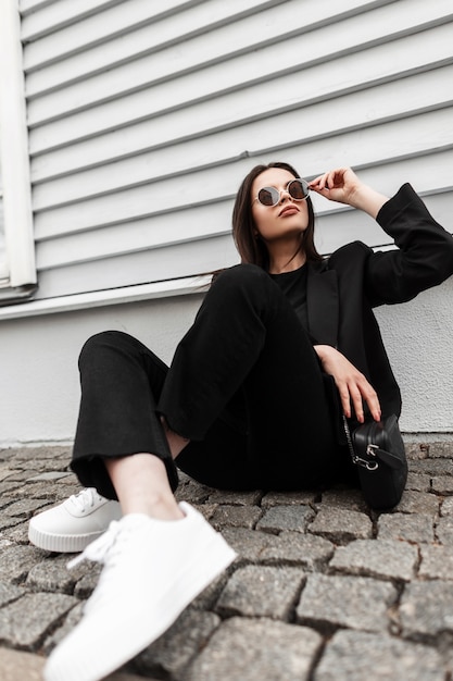 Pretty young business woman fashion model in white shoes in stylish black clothing with handbag straightens fashion sunglasses near wooden wall in city. Beaty girl in casual wear sit on stone tile.