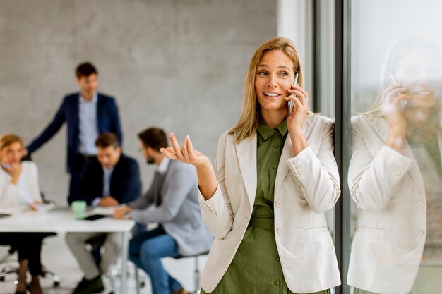 Pretty young business woman standing in the office and using mobile phone in front of her team