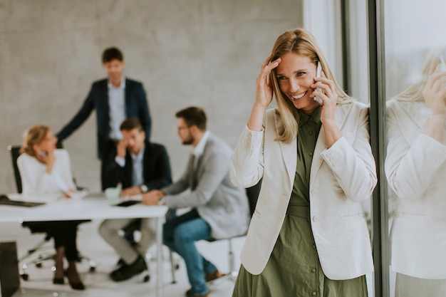 Pretty young business woman standing in the office and using mobile phone in front of her team