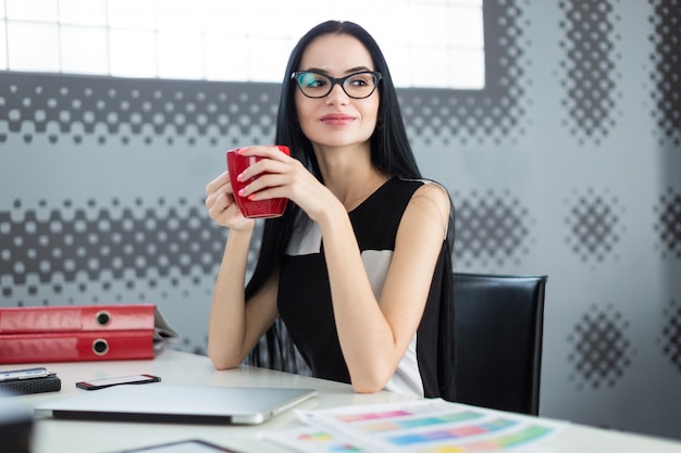 Pretty, young businesslady in black dress and glasses sit at the table and hold red cup