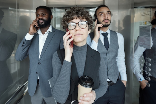 Pretty young businesswoman and her two intercultural colleagues with smartphones by ears moving by elevator for work