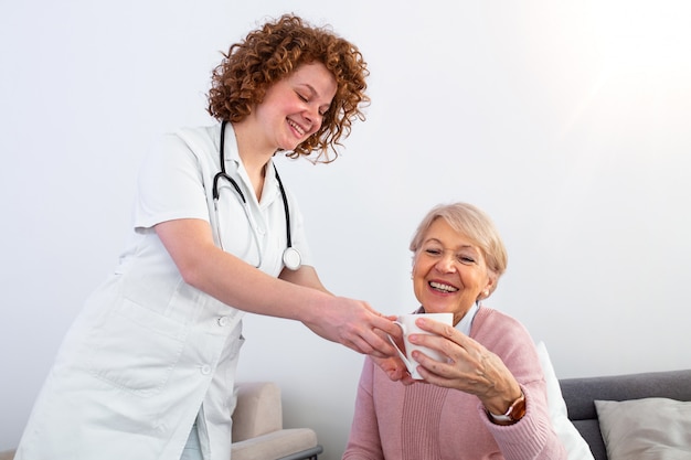 Pretty young caregiver serving afternoon cup of tea to older happy woman. Young nurse caring for elderly patient in her home.