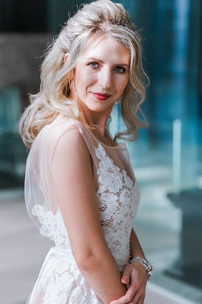 Pretty young caucasian blonde bride with makeup and trendy hairstyle in wedding dress standing and posing in interior