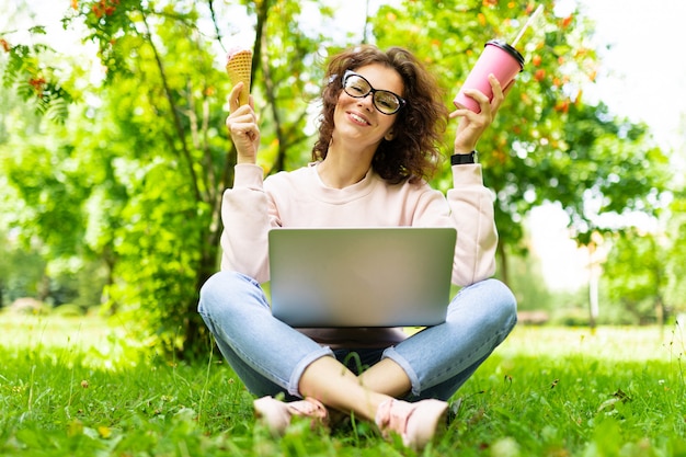 Pretty young caucasian woman is engaged in freelance in the park with the cup of coffe and ice-cream