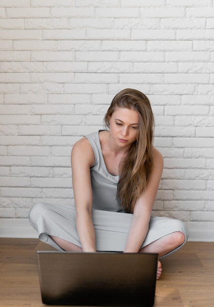 Pretty young caucasian woman sitting on the floor while typing on laptop on white brick background. Communicaton, technology concept