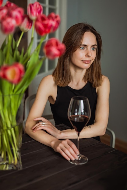 Una bella giovane donna caucasica seduta al tavolo di legno in cucina con un bicchiere di vino rosso in mano un bouquet di tulipani