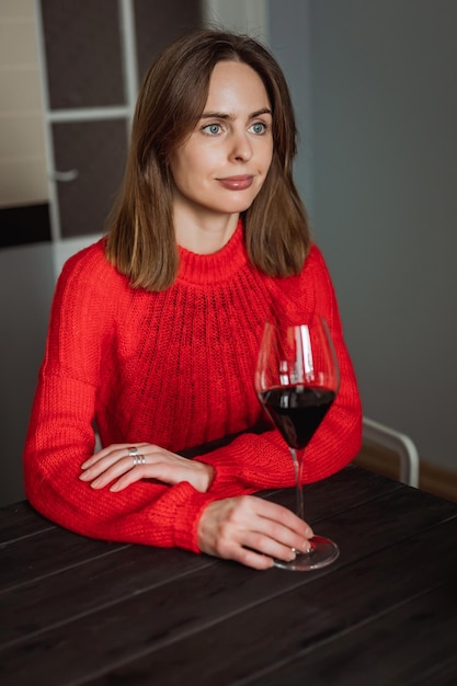 Una bella giovane donna caucasica seduta al tavolo di legno in cucina con un bicchiere di vino rosso in mano