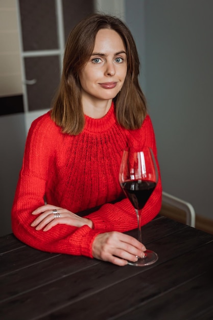 A pretty young caucasian woman sitting at the wooden table in the kitchen with a glass of red wine in her hands