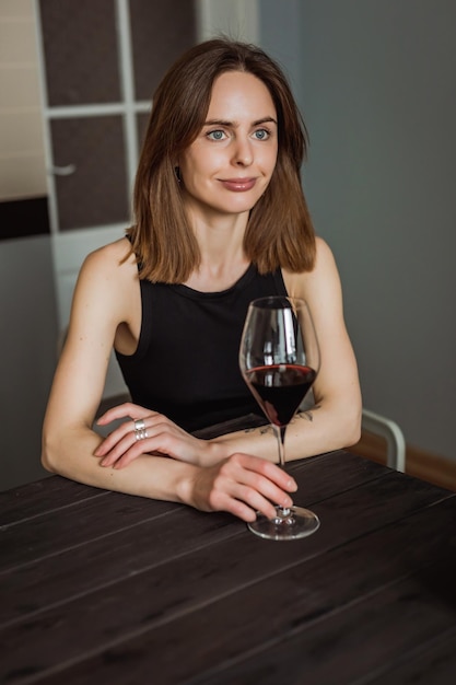 彼女の手に赤ワインのグラスを持って台所の木製のテーブルに座っているかなり若い白人女性