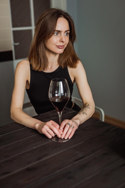 A pretty young caucasian woman sitting at the wooden table in the kitchen with a glass of red wine in her hands