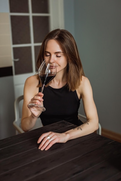 A pretty young caucasian woman tasting a glass of red wine sitting at the wooden table in the kitchen