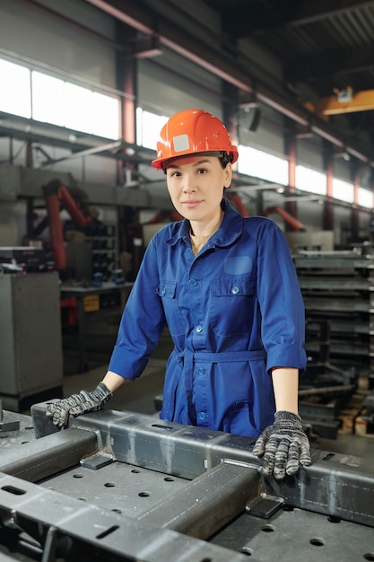 Pretty young confident factory worker in blue uniform, gloves and helmet posing