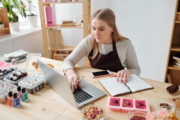 Piuttosto giovane donna creativa in grembiule seduto dal posto di lavoro davanti al computer portatile mentre prende appunti sul suo hobby