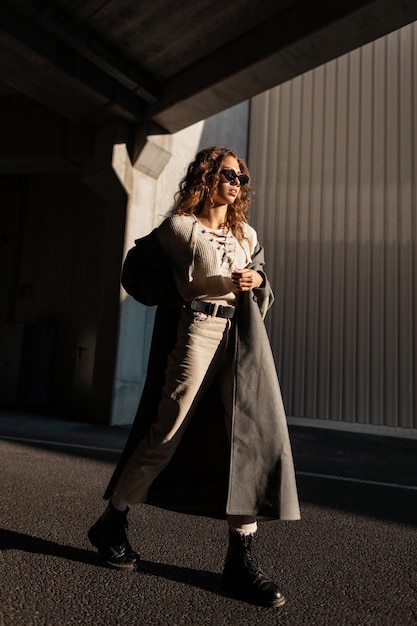 La ragazza riccia abbastanza giovane con gli occhiali da sole in vestiti alla moda sembra indossare un cappotto lungo passeggiate in città alla luce del sole. stile e bellezza femminili urbani