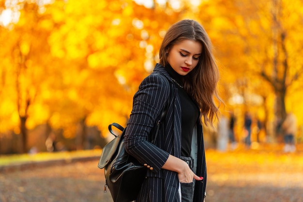 Pretty young elegant business woman with a black suit in a jacket with a leather backpack walks in an autumn park with bright colored orange foliage at sunset