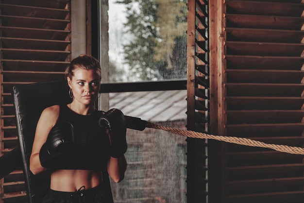 かなり若い女性のボクサーは、ボクシンググローブをはめて、古いジムのサンドバッグでリングのトレーニングをしています。ボックストレーニングの女性。健康的なライフスタイル、スポーツ、ジムでの運動の概念。コピースペース