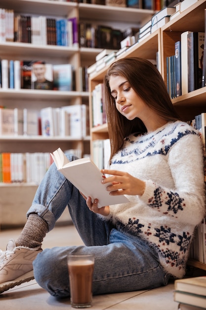 Pretty young female student reading book on library floor at the university