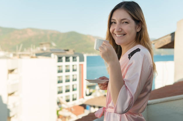 Pretty young girl in bathrobe drinking coffee while standing on the balcony of her room at the hotel and looking at camera