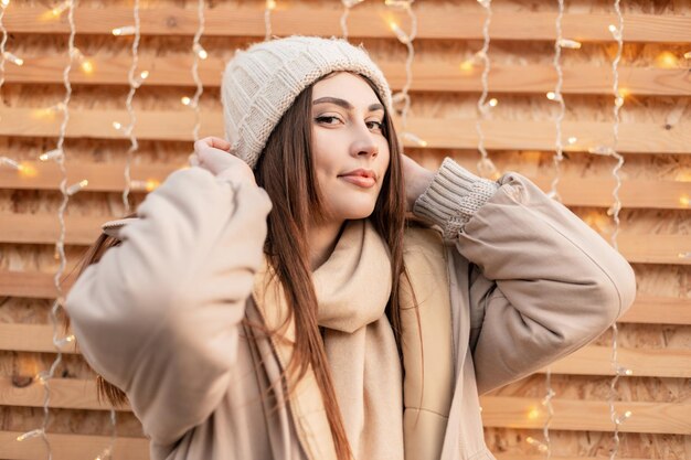 Pretty young girl in fashionable winter outwear with a knitted vintage slipper, a fashion Down-padded coat and a scarf stands near a wooden wall with lights