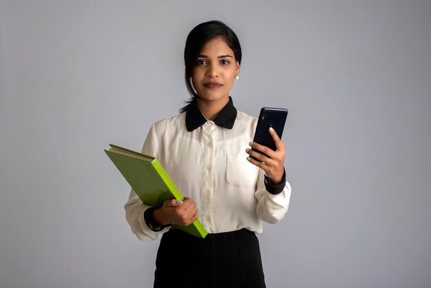 Pretty young girl holding book and using mobile on grey wall