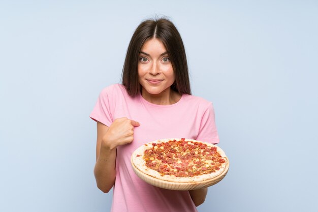 Pretty young girl holding a pizza over isolated blue wall with surprise facial expression