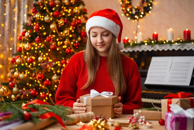 Красивая молодая девушка с подарочной коробкой сидит в красивой комнате, полной огней и рождественских украшений. Зимние каникулы. Время волшебства в воздухе.