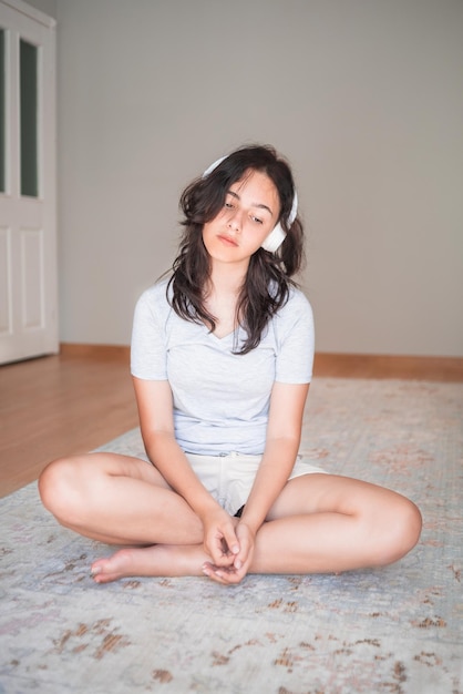 Bella ragazza che ascolta la musica con le cuffie mentre si siede sul tappeto a casa appartamento che vive vita domestica