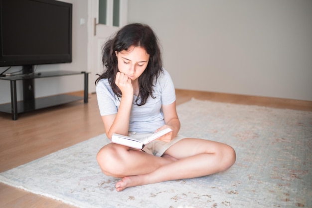 Bella ragazza che legge il libro di carta mentre si siede sul tappeto a casa