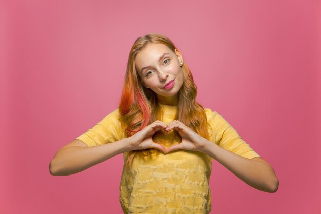 Pretty young girl show heart gesture with love feeling romantic mood sympathy support on pink studio