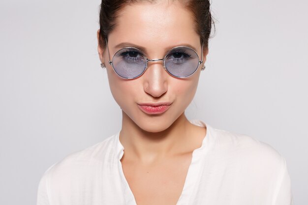 Pretty young girl in white blank t-shirt, wearing glasses, empty wall, studio portrait. Smiling brunette woman in eyeglasses