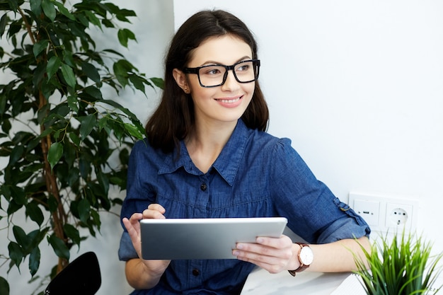 Pretty young  girl with black hair wearing blue stripped shirt and eyeglasses sitting in cafe with cup of coffee, freelance concept, portrait, smiling, holding tablet and looking right.