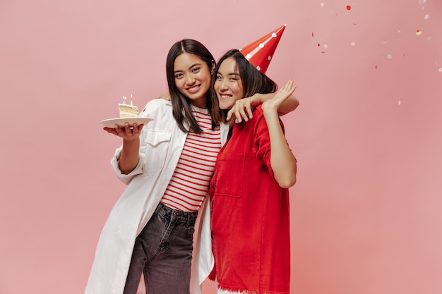 Pretty young girls celebrate birthday and smile on pink isolated background Brunette woman holds piece of cake and hugs her friend in party hat