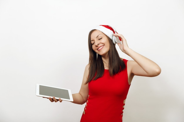 Pretty young happy woman in red dress and Christmas hat listening music on headphones, holding tablet on white background. Santa girl gadget isolated. New Year holiday 2018. Copy space advertisement.