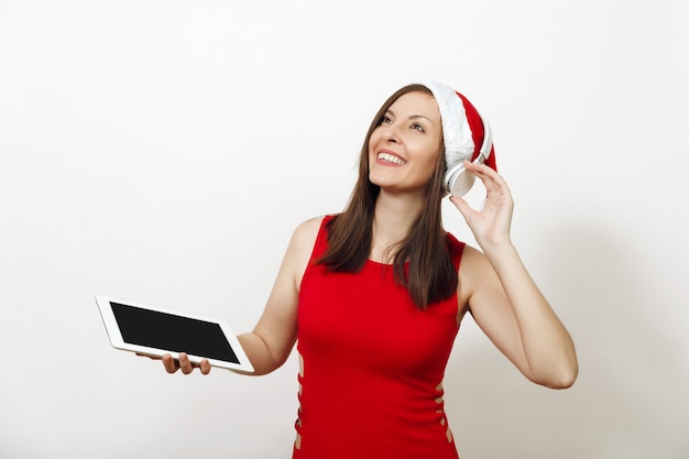 Довольно молодая счастливая женщина в красном платье и рождественская шляпа слушает музыку на наушниках, держа таблетку на белом фоне. Изолированный гаджет девушки Санты. Новый год праздник 2018. Копирование пространства рекламы.