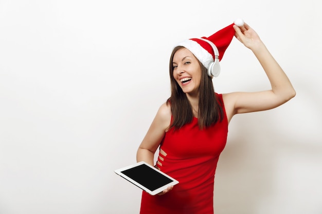 Pretty young happy woman in red dress and Christmas hat listening music on headphones, holding tablet on white background. Santa girl gadget isolated. New Year holiday 2018. Copy space advertisement.