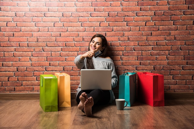 Pretty young Indian woman or girl shopping with debit or Credit card and laptop while sitting over wooden floor against red brick wall