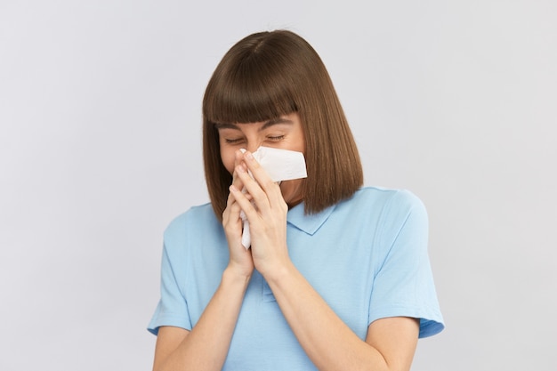 Pretty young lady hardly sneezing her nose into tissue in blue shirt on grey wall