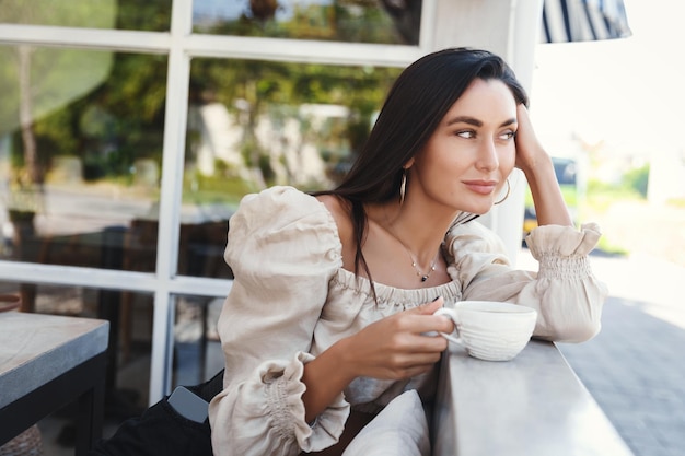 エレガントなブラウスに座って屋外カフェでコーヒーを飲み、晴れた夏の日にレストランで通りの女性を見ている黄金色の日焼けのかなり若い女性