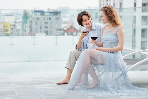 Pretty young lesbian women sitting on rooftop, talking and drinking tasty red wine