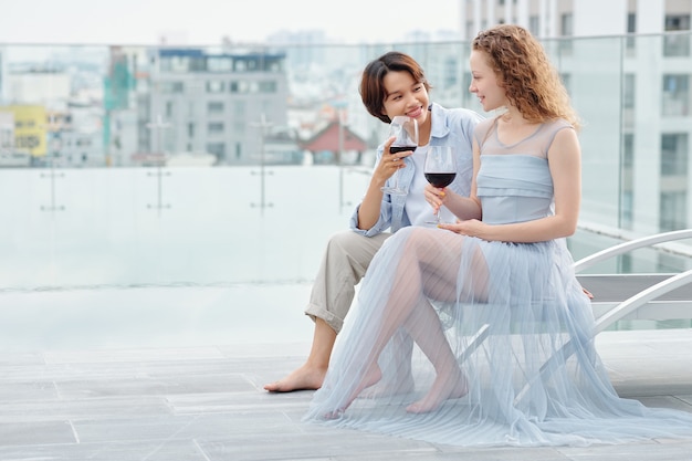 Photo pretty young lesbian women sitting on rooftop, talking and drinking tasty red wine