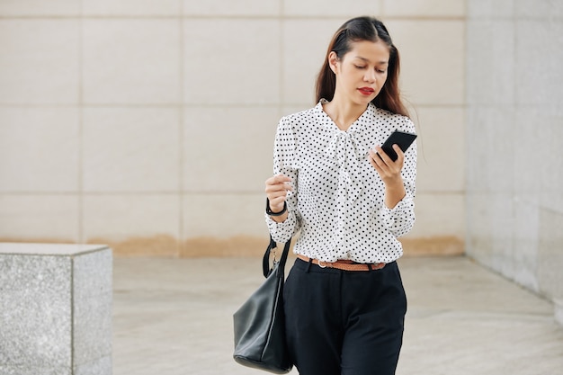 Piuttosto giovane giovane imprenditrice multietnica in camicetta a pois camminare all'aperto anc leggendo il messaggio di testo sullo smartphone