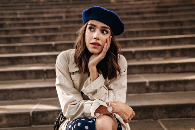 Pretty young Parisienne in blue beret, beige trench coat and polka dot skirt, sitting on stairs outdoors and anxiously looking away