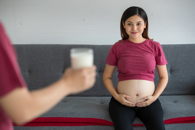 소파에 앉아 음료수 한 잔을 바라보며 웃고 있는 빨간 티셔츠를 입은 꽤 젊은 임신한 아시아 여성. 우유 한 잔을 들고 손에 부드러운 초점. 임신 건강 개념입니다.