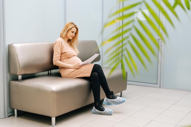 Pretty young pregnant woman is sitting in hallway of hospital waiting to see gynecologist Expectant mother studying medical records and examination results Concept of happy and healthy childbearing