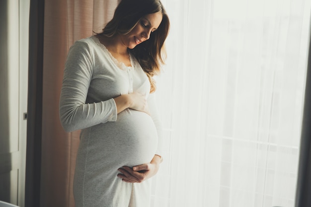 Pretty young pregnant woman standing by the window in room