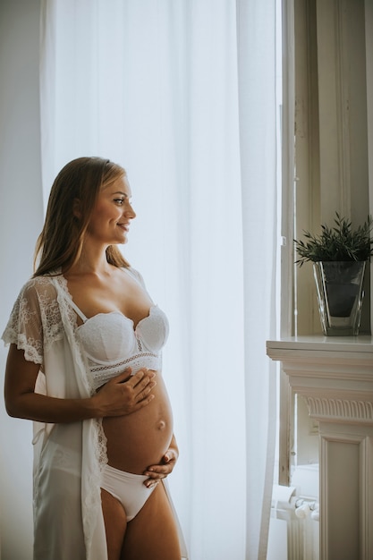 Pretty young pregnant woman standing at window in the room at daylight