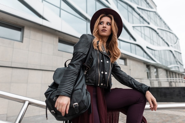 Pretty young red-haired woman in a purple vintage hat in a black stylish leather jacket with a fashionable backpack in trendy pants