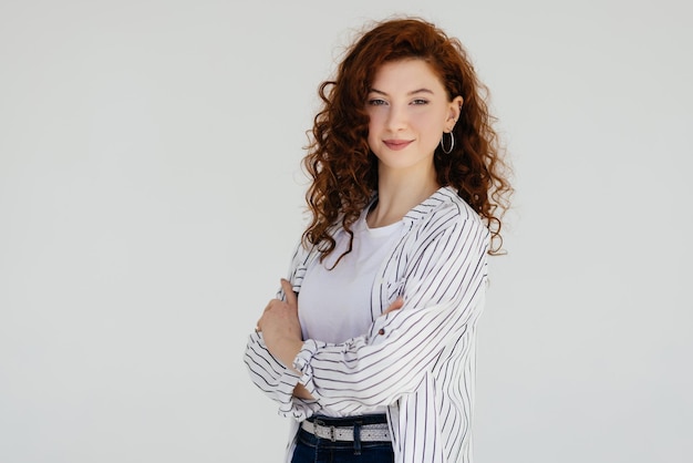 Pretty young redhead lady standing isolated over white wall background with arms crossed